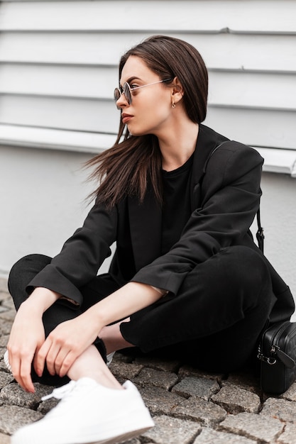 Mannequin urbaine de jeune femme dans des lunettes de soleil à la mode dans des chaussures blanches dans des vêtements noirs élégants avec un sac à main se détendre près d'un bâtiment en bois en ville. Jolie fille en tenue décontractée se repose à l'extérieur.