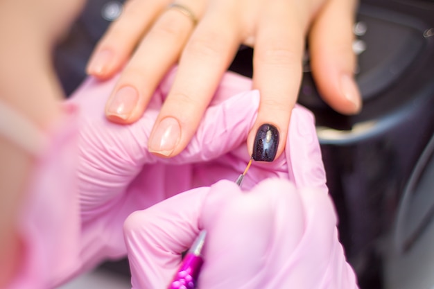 Manucure. Closeup Manicurist les mains dans des gants roses est un vernis à ongles noir sur les ongles du client.