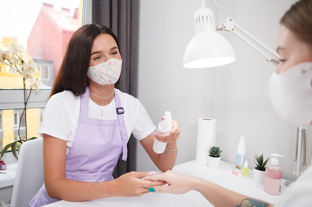 Manucure professionnelle portant un masque facial, désinfectant les mains d'une cliente