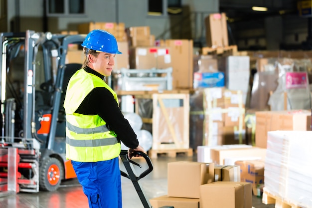 Un manutentionnaire en veste de protection tire un déménageur avec des colis et des boîtes à l'entrepôt d'une entreprise de transport