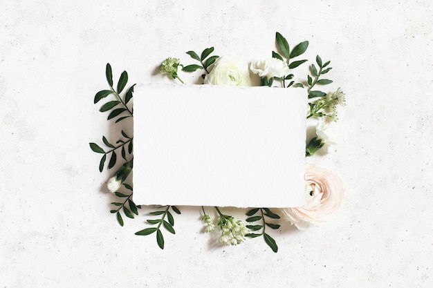 Photo maquette d'anniversaire de mariage féminin carte de voeux en papier vierge cadre floral de renoncule rose blanc oeillet et fleurs d'astrantia feuilles de lentisque fond de table en béton vue de dessus à plat