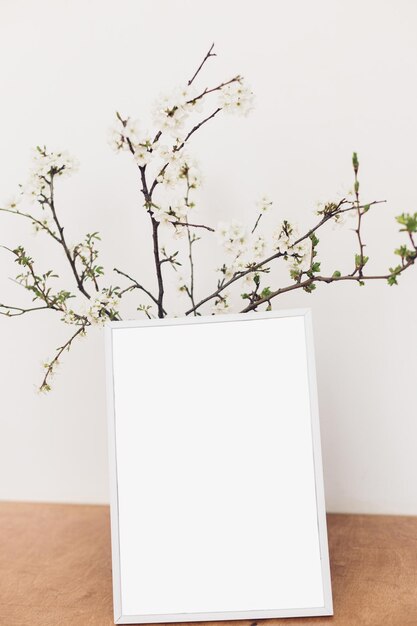 Maquette de cadre photo et branches de cerisier en fleurs sur une table en bois contre un mur blanc Modèle de cadre photo vide et fleurs de printemps Vie à la campagne simple Espace pour le texte