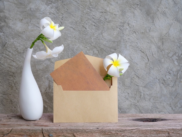 Maquette carte brune d'invitation et belles fleurs de Plumeria dans un vase blanc moderne sur une table en bois grunge avec fond de mur loft, carte de voeux dans un ton doux nature morte