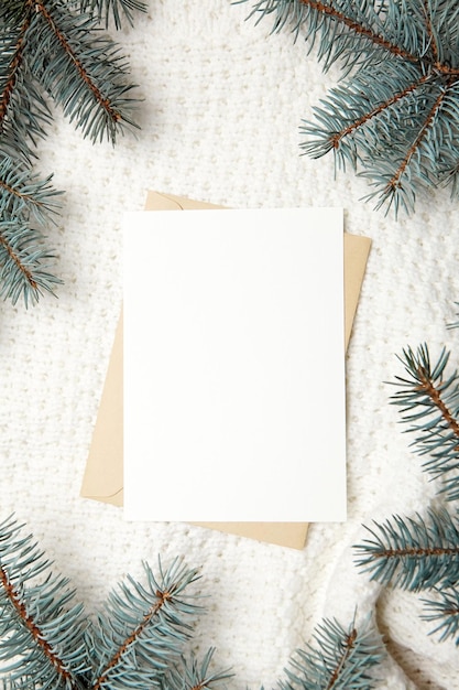 Maquette de carte de voeux de noël avec enveloppe et branches de sapin sur la vue de dessus de fond de pull tricoté d'hiver blanc