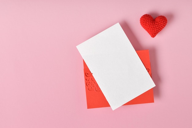 Une maquette d'une enveloppe rouge avec une feuille vierge et un coeur tricoté rouge sur fond rose