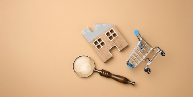 Maquette d'une maison en bois, d'un caddie miniature et d'une loupe sur fond marron clair. Concept de recherche de maison à louer, achat, hypothèque