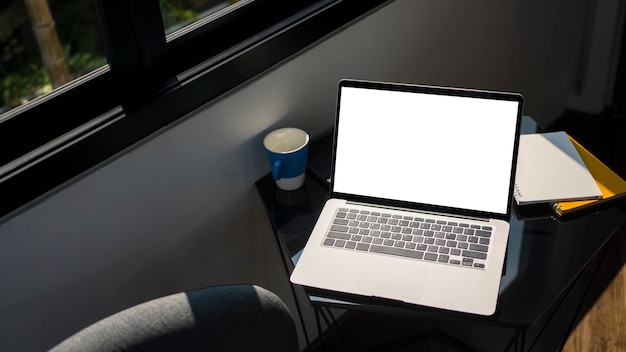 Maquette d'ordinateur portable avec écran blanc, paperasse et tasse à café sur une table noire au bureau.