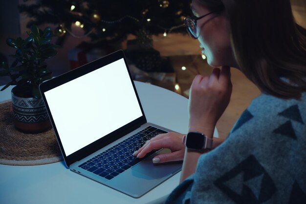 Maquette d'ordinateur portable à écran blanc vierge femme utilisant un ordinateur portable et une montre intelligente à la maison sur fond d'arbre de noël et de lumières