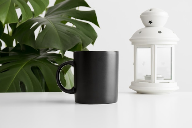 Maquette de tasse noire avec un bougeoir sur un tableau blanc et une plante monstera