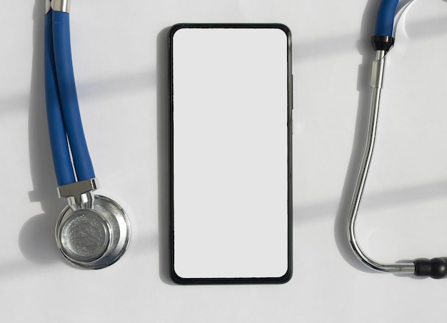 Maquette de téléphone portable avec stéthoscope sur un bureau blanc, vue de dessus. Modèle avec écran blanc de smartphone pour application médicale.