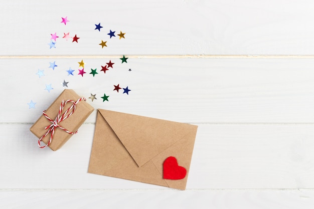 Photo maquette de vacances: coffrets cadeaux, coeur rouge et papier vierge dans une enveloppe brune sur un fond en bois blanc