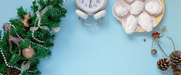 Maquette vue de dessus de carte de voeux de Noël et arbre du nouvel an, plat sur fond bleu avec des pommes de sapin, horloge, biscuits, place pour votre texte sur une feuille blanche au milieu.