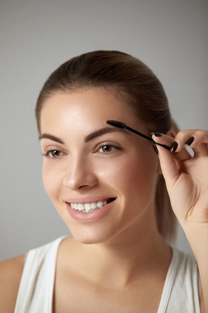 Maquillage de beauté. Belle femme façonnant le gros plan des sourcils. Modèle de fille avec un maquillage professionnel à l'aide d'une brosse à sourcils et souriant