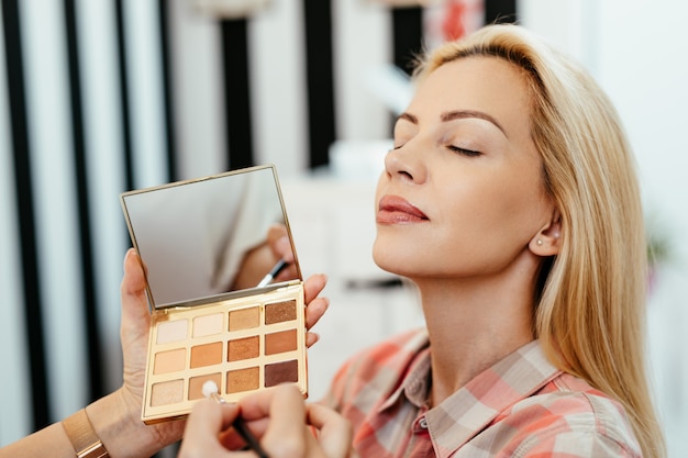 Maquilleuse appliquant le maquillage professionnel d'une belle femme blonde d'âge moyen. Gros plan sur la palette de fards à paupières.