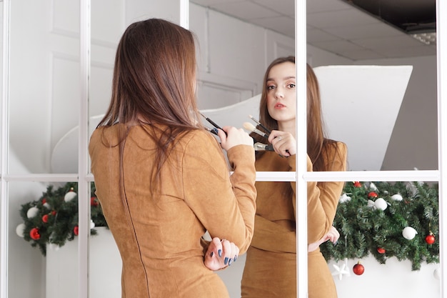 Maquilleuse aux longs cheveux noirs tenant un pinceau à poudre, expression hautaine, reflet dans le miroir, décor de Noël derrière