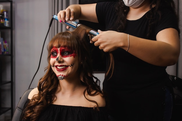 Maquilleuse frisant les cheveux d'une fille souriante avec du maquillage Dia de los Muertos.