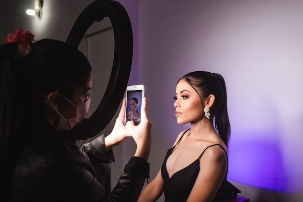 Maquilleuse prenant des photos avec un anneau lumineux à une belle fille avec un maquillage artistique sur un mur blanc avec des lumières.
