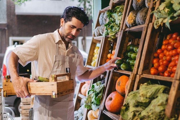 Marchand de légumes latin d'âge moyen avec un tablier et une caisse à la main organisant les légumes dans son magasin