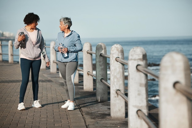 Marche sportive et femmes au bord de l'océan parlant pour un mode de vie sain, bien-être et cardio sur la promenade Amis sportifs et femmes en conversation sur la promenade pour l'entraînement physique et l'entraînement