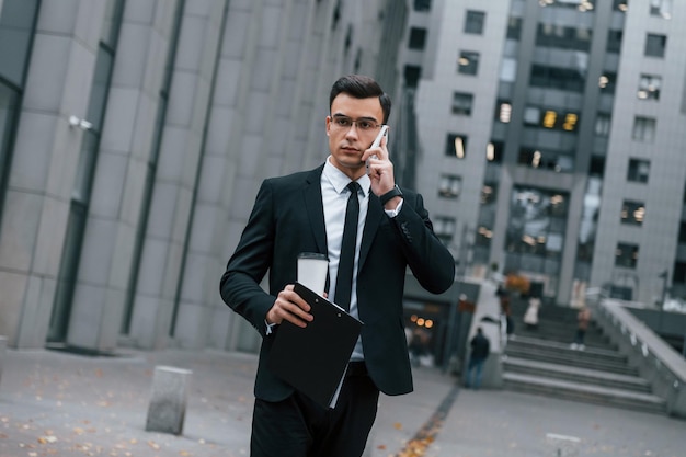 Marcher et parler par téléphone Homme d'affaires en costume noir et cravate est à l'extérieur de la ville