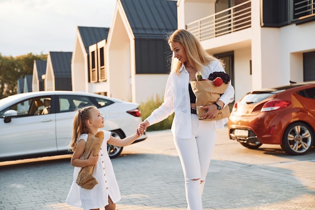 Marcher et tenir des sacs avec des produits Jeune femme avec sa petite fille est avec leur voiture électrique à l'extérieur