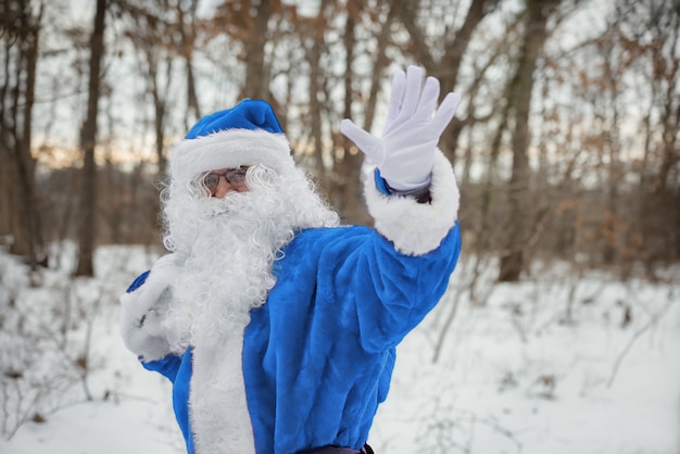 Marcher à travers la forêt d'hiver, agite sa main Père Noël en costume bleu transportant des cadeaux de Noël