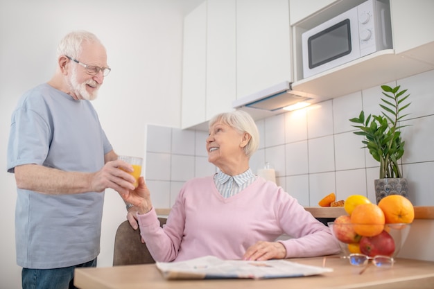 Mari attentionné. Senior homme aux cheveux gris donnant un verre de jus d'orange à sa femme