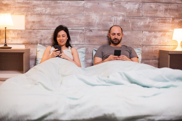 Mari ennuyé naviguant sur smartphone avant le coucher. Belle femme en pyjama souriante.