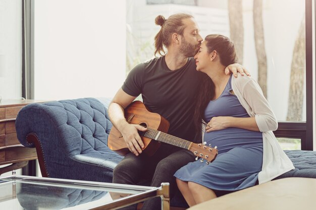 Le mari avec la femme enceinte reste à la maison en jouant de la guitare pour bébé qui s'embrasse et montre de l'amour ensemble