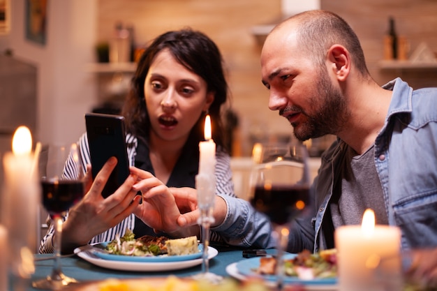 Le mari et la femme ont l'air choqués au téléphone pendant un dîner romantique dans la cuisine Des adultes assis à table dans la cuisine naviguent, cherchent, utilisent des smartphones, Internet, célèbrent leur anniversaire.