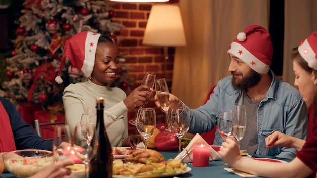 Mari festif parlant avec une femme afro-américaine tout en faisant tinter des verres de champagne au dîner de Noël. Joyeuses personnes diverses portant des chapeaux traditionnels célébrant les vacances d'hiver.