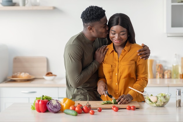 Un mari noir aimant embrasse sa femme pendant qu'elle cuisine des aliments à l'intérieur de la cuisine