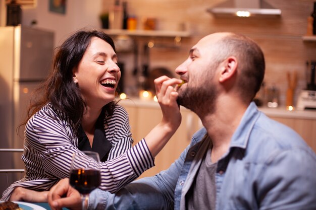 Mari nourrissant sa femme avec des raisins lors d'un dîner romantique célébrant la relation. L'épouse et le mari célèbrent leur anniversaire avec du vin rouge, des moments tendres aux chandelles dans la cuisine.