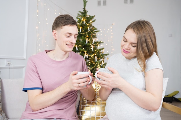 Mari prenant soin de sa femme enceinte en donnant une tasse de thé sur fond d'arbre de Noël.