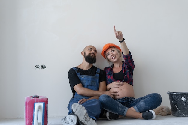 Le mari et sa femme enceinte sont assis par terre dans la pièce. uniforme de travail et casque de sécurité orange. pointez vers le haut et regardez l'espace libre pour le texte.