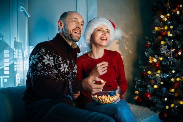 Un mari et sa femme savourent du pop-corn la veille de Noël en regardant la télévision et en riant fort