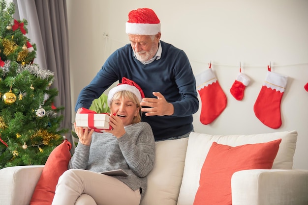 Le mari Senier donne une boîte-cadeau à sa femme Assis sur le canapé et portant un chapeau de Père Noël