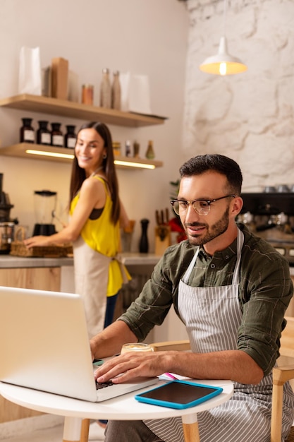 Mari travaillant sur un ordinateur portable pendant que sa femme prépare du café dans leur cafétéria