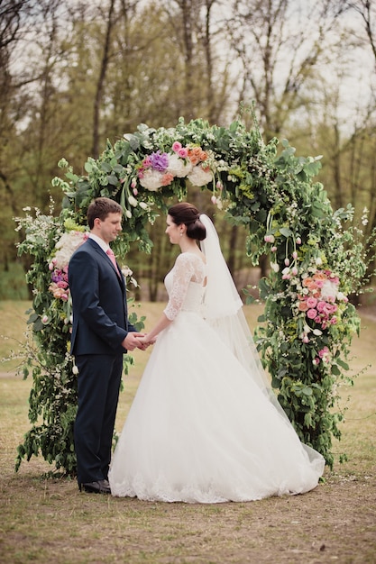 Mariage au printemps. Cérémonie dans la rue. Une arche de vraies fleurs. La mariée et le marié se regardent.