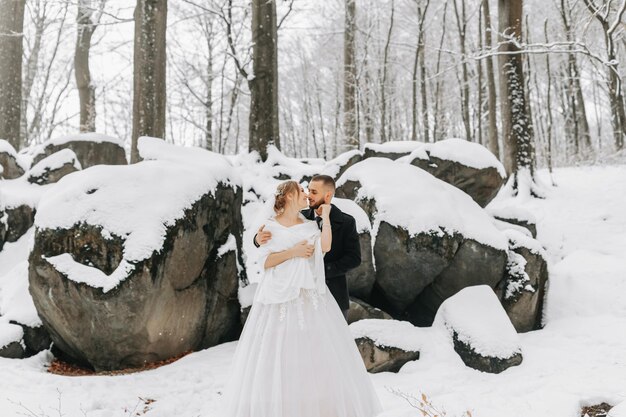 Le mariage en hiver La mariée et le marié se tiennent sur le fond de rochers couverts de neige La mariée dans une robe blanche et un poncho blanc Le marié dans un manteau noir