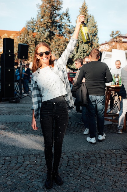 Maribor, Slovénie - 11 Octobre 2019 : Une Jeune Fille Souriante Portant Des Lunettes De Soleil Tient Une Bouteille De Vin. Style Décontracté Femme. Personne De Sexe Féminin S'amusant. Joyeuses Fêtes. Portrait De Style De Vie En Plein Air.