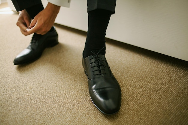 Le marié assis sur le canapé et attaché des lacets sur des chaussures en cuir noir, gros plan. mariage