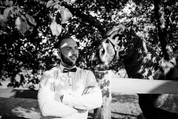 Le marié en chemise blanche et nœud papillon violet. Équitation. Photographie en noir et blanc.