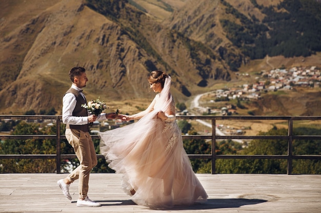 Marié en costume moderne et mariée en charmante robe rose pose sur la terasse avec vue sur la montagne en Géorgie