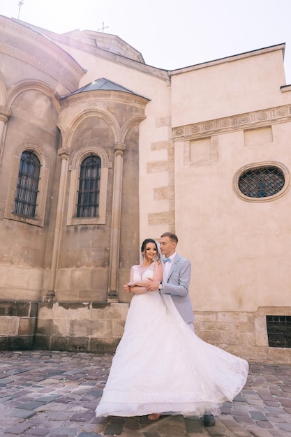 Un marié élégant en costume et une belle mariée en robe blanche s'étreignent sur le fond du bâtiment Portrait de mariage de jeunes mariés amoureux