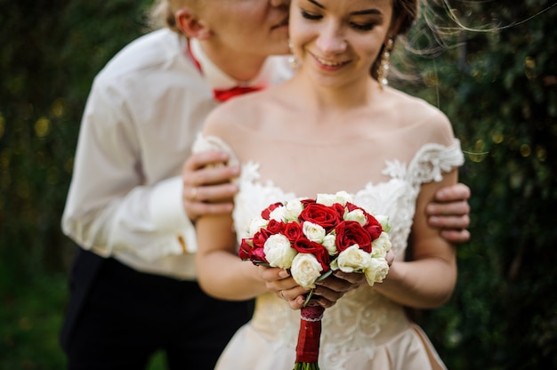 Marié embrassant sa jeune et belle mariée qui tient un bouquet de mariage en arrière-plan de l'arbre vert