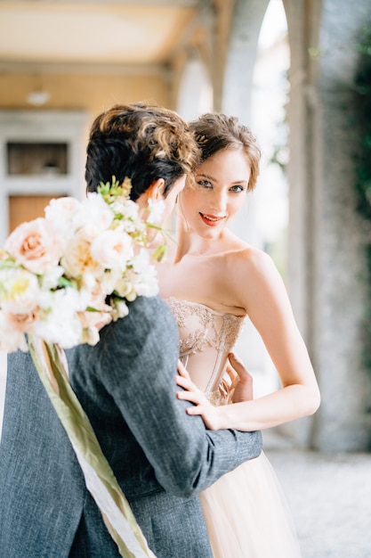 Le marié embrasse la mariée avec un bouquet de fleurs sur une ancienne terrasse avec des arcs enlacés de lierre vert