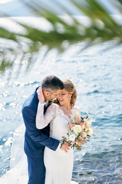 Photo le marié embrasse la mariée souriante avec un bouquet de derrière près de la mer brillante