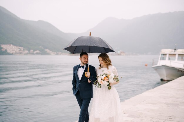 Le marié et la mariée sous un parapluie marchent le long de la jetée