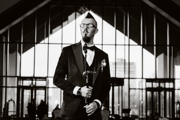 Le marié en smoking et nœud papillon avec une coiffure mohawk et une canne à l'intérieur